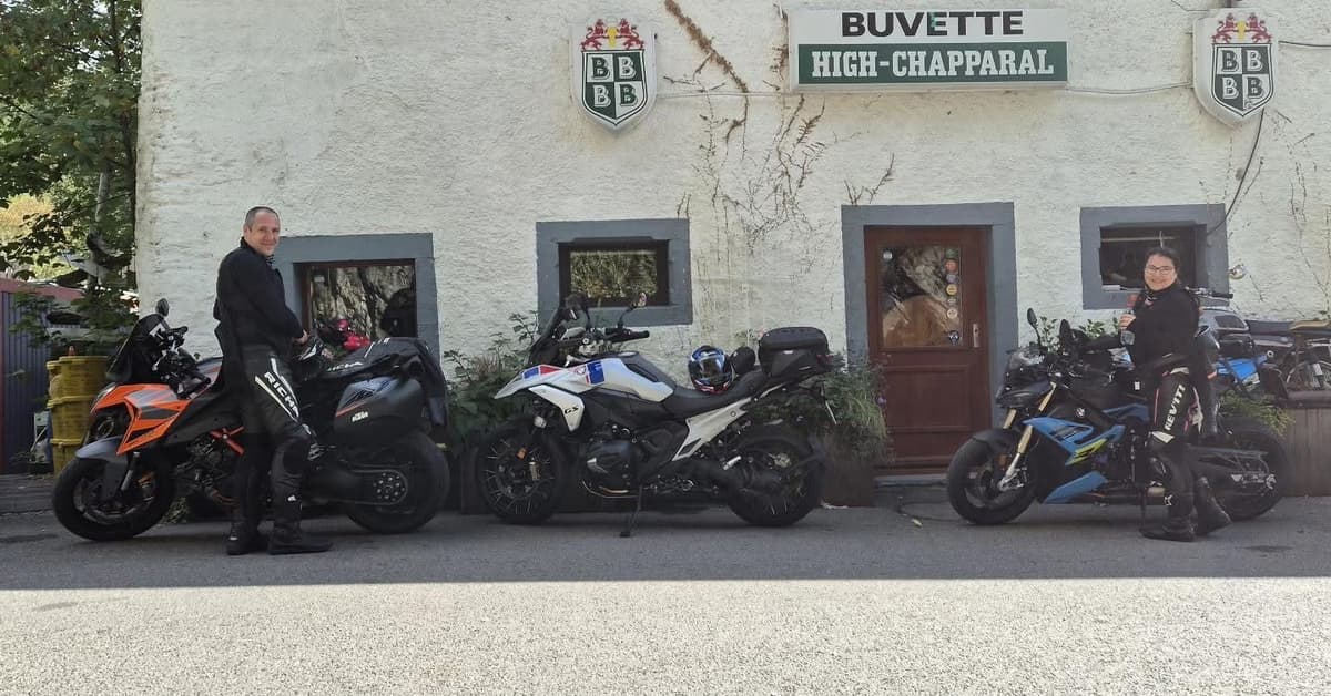 Twee motorrijders bij hun motoren, een oranje KTM, een witte BMW GS en een blauwe naked bike, voor de witte gevel van Buvette High-Chapparal.