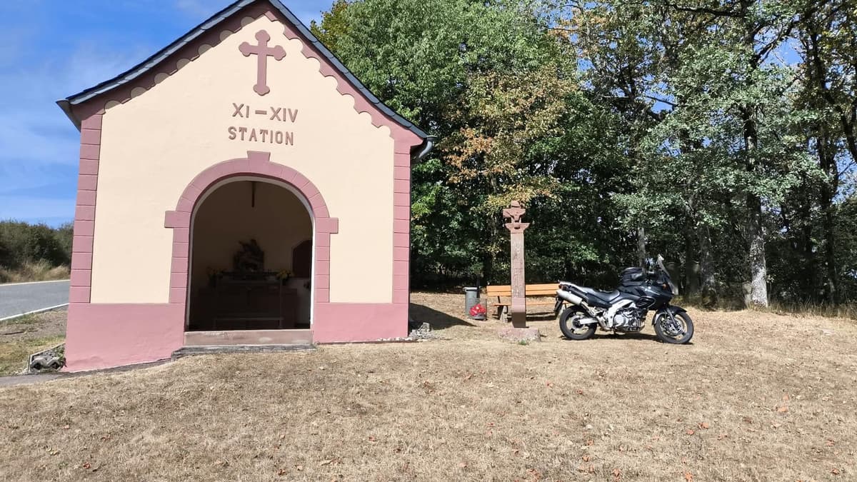 Een roze-wit kapelletje met een kruis langs de weg, met ernaast een stenen kruis, een bankje en een geparkeerde motor in het gras.