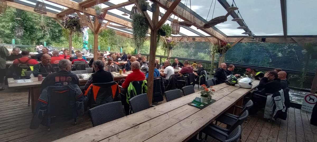 Een groot overdekt houten terras vol motorrijders aan lange tafels, met bomen en water op de achtergrond.