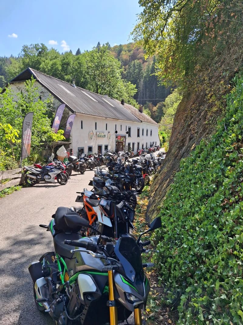 Een lange rij motoren langs een smalle weg tussen een met klimop begroeide rotswand en een wit gebouw met het opschrift Buvette High-Chapparal.