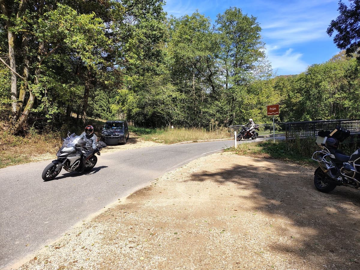 Een motorrijder in een grijs pak neemt een bocht op een smal baantje langs het bos, met verderop een tweede rijder bij de brug over de Prüm.