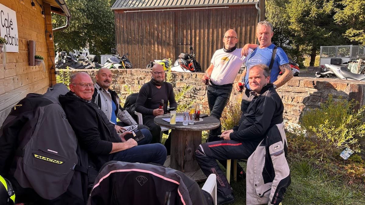 Zes motorrijders in de avondzon rond een tafel van een kabelhaspel, in een tuintje achter een stenen muur met hun motoren erachter.