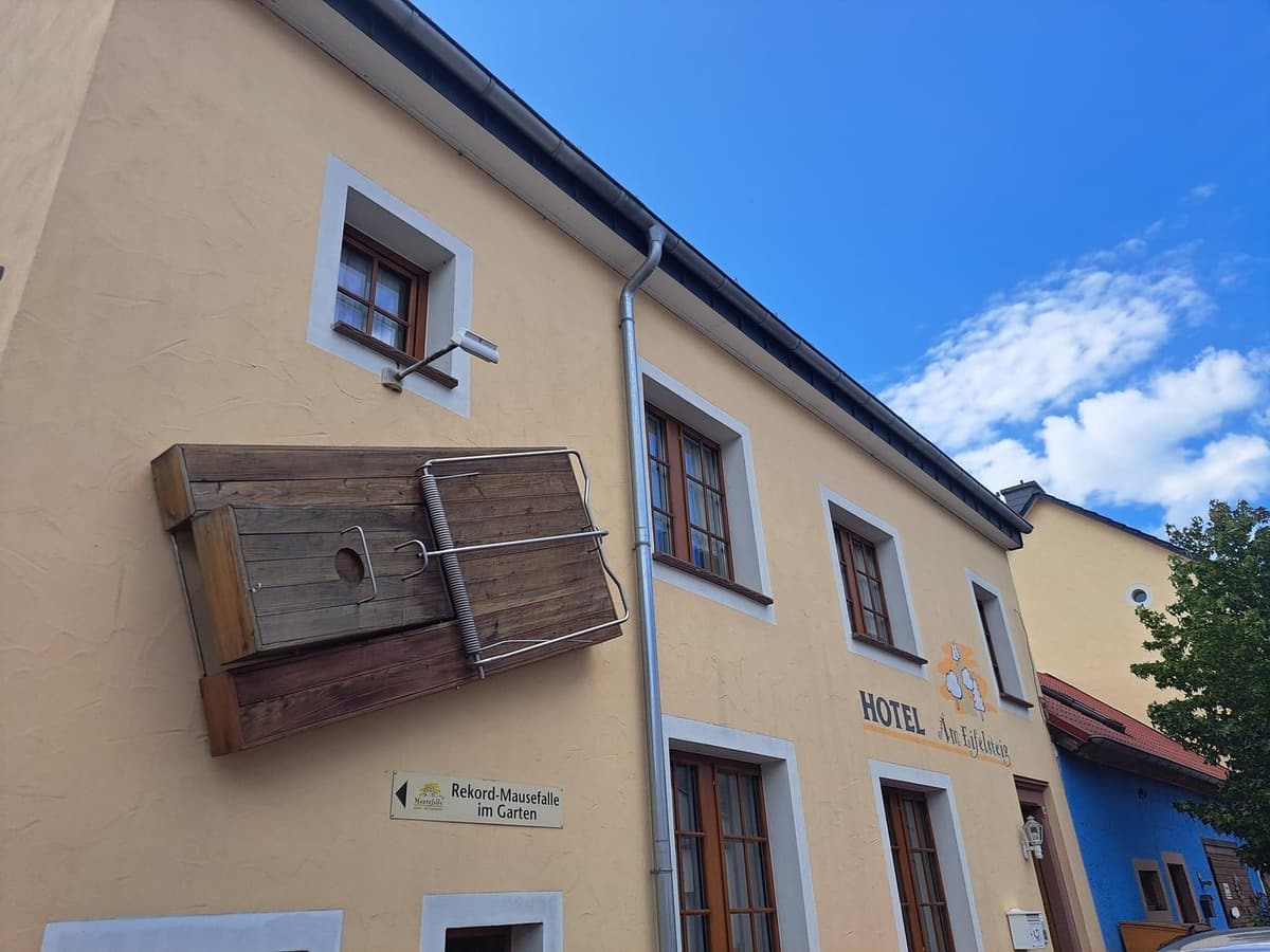 De gele gevel van Hotel Am Eifelsteig in Neroth, met een grote houten muizenval aan de muur en een bordje naar de Rekord-Mausefalle in de tuin.