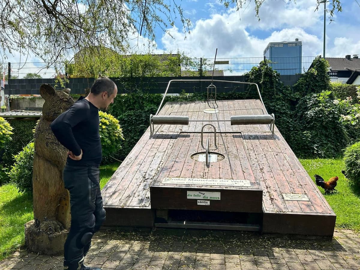 Een motorrijder bekijkt een reusachtige houten muizenval in een tuin, met ernaast een houten beer en een haan in het gras.