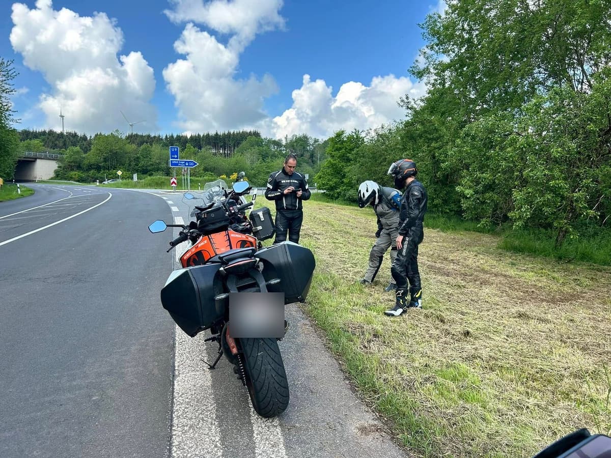 Een oranje-zwarte motor met koffers in de berm van een bochtige weg, met drie motorrijders in het gras en een wegwijzer naar Prüm.