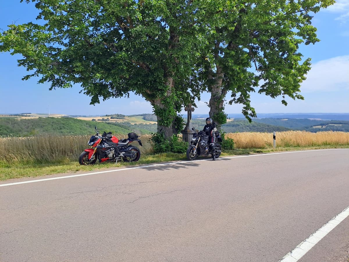 Een rode en een zwarte motor onder twee grote bomen bij een stenen wegkruis, met een rijder op de zwarte motor en daarachter graanvelden en heuvels.