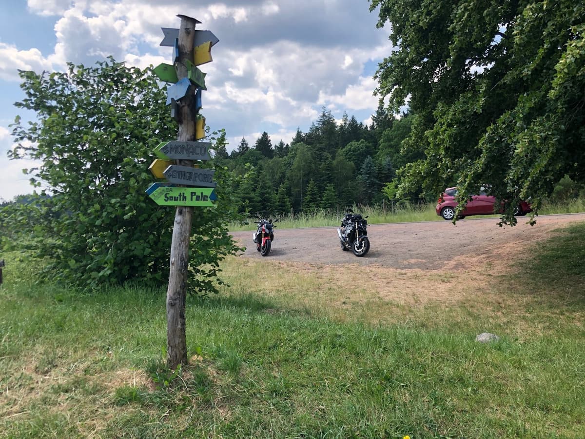 Een houten paal vol gekleurde wegwijzers naar Monaco, Singapore en de South Pole, met twee motoren op een parking aan de rand van het bos.