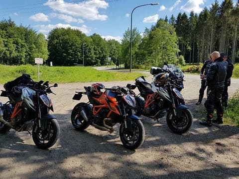 Drie oranje KTM-naked bikes op een grindstrook langs een bos, met hun rijders ernaast.