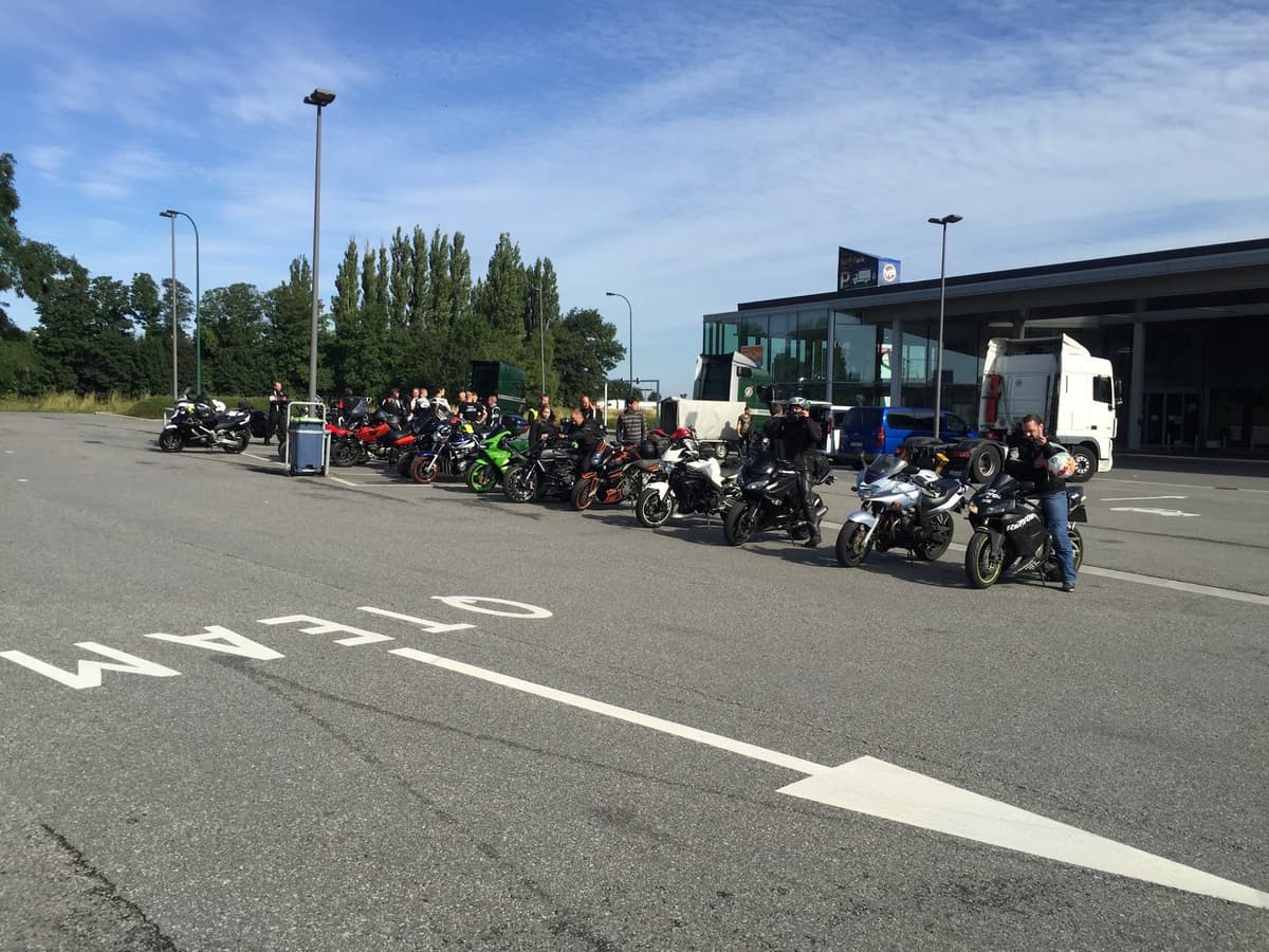 Een rij motoren en motorrijders op een parking naast een tankstation, met witte pijlen op het asfalt.