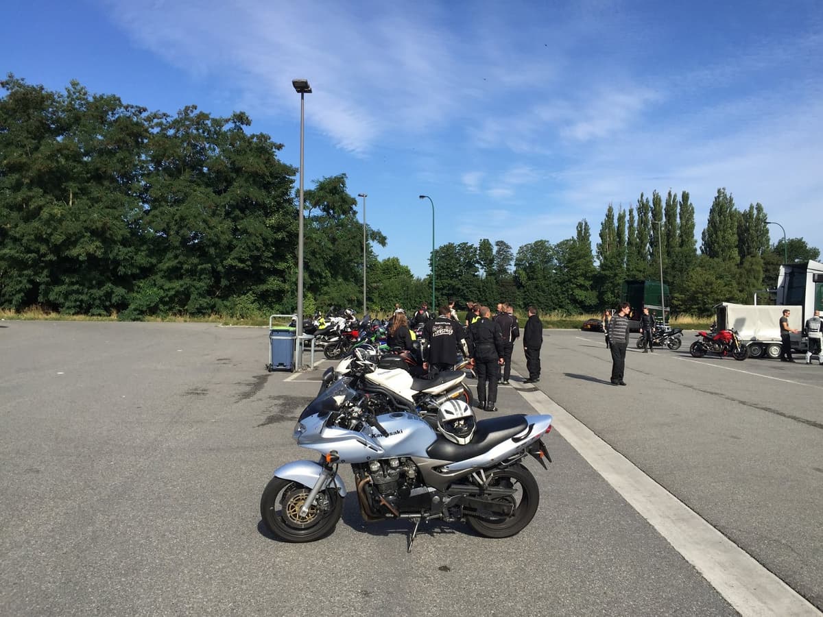 Een zilveren Kawasaki op een grote parking, met daarachter een groep motorrijders tussen hun motoren onder een blauwe lucht.
