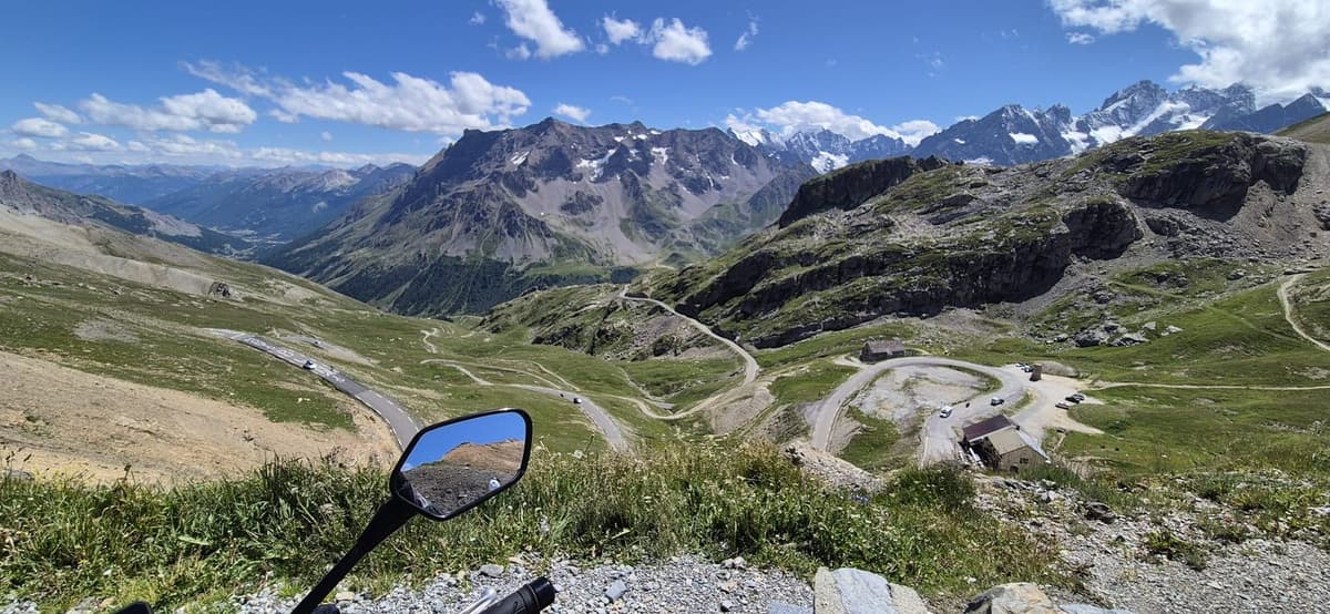 Zicht vanaf een col op een bergweg met haarspeldbochten door groene alpenweiden, met besneeuwde toppen aan de horizon en vooraan de spiegel van een motor.