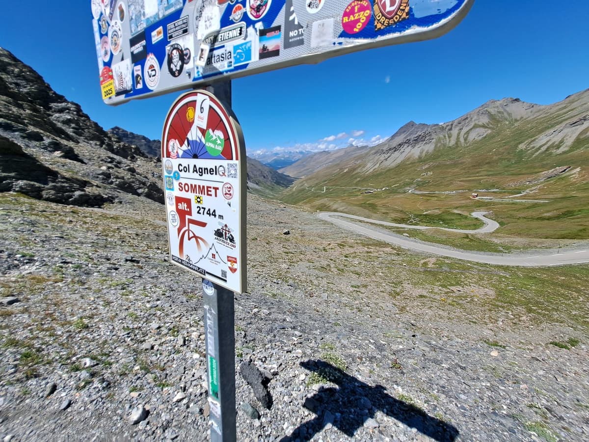 Het bord op de top van de Col Agnel, 2.744 meter, vol stickers, met daarachter een bochtige weg door een kale bergvallei.