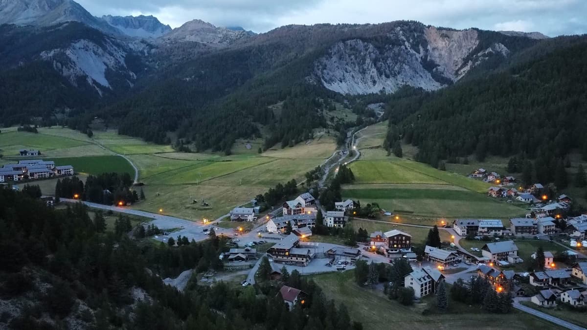 Zicht van bovenaf op een bergdorp in de schemering, met brandende straatlantaarns, weiden en beboste hellingen.