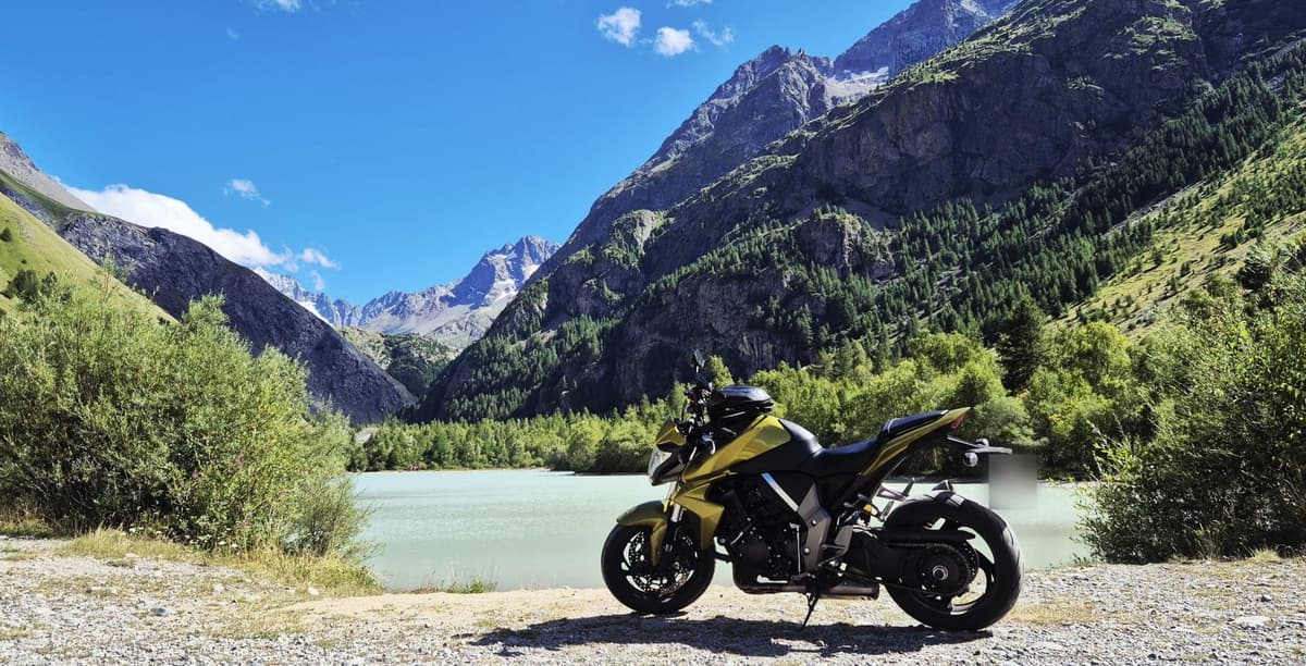 Een gele naked bike staat aan een lichtblauw bergmeer, tussen beboste hellingen en besneeuwde toppen.