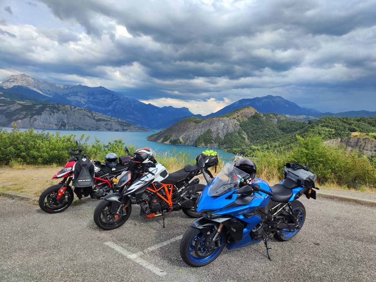 Drie motoren staan op een parking boven een turquoise meer met een beboste landtong, onder een donkere wolkenlucht.