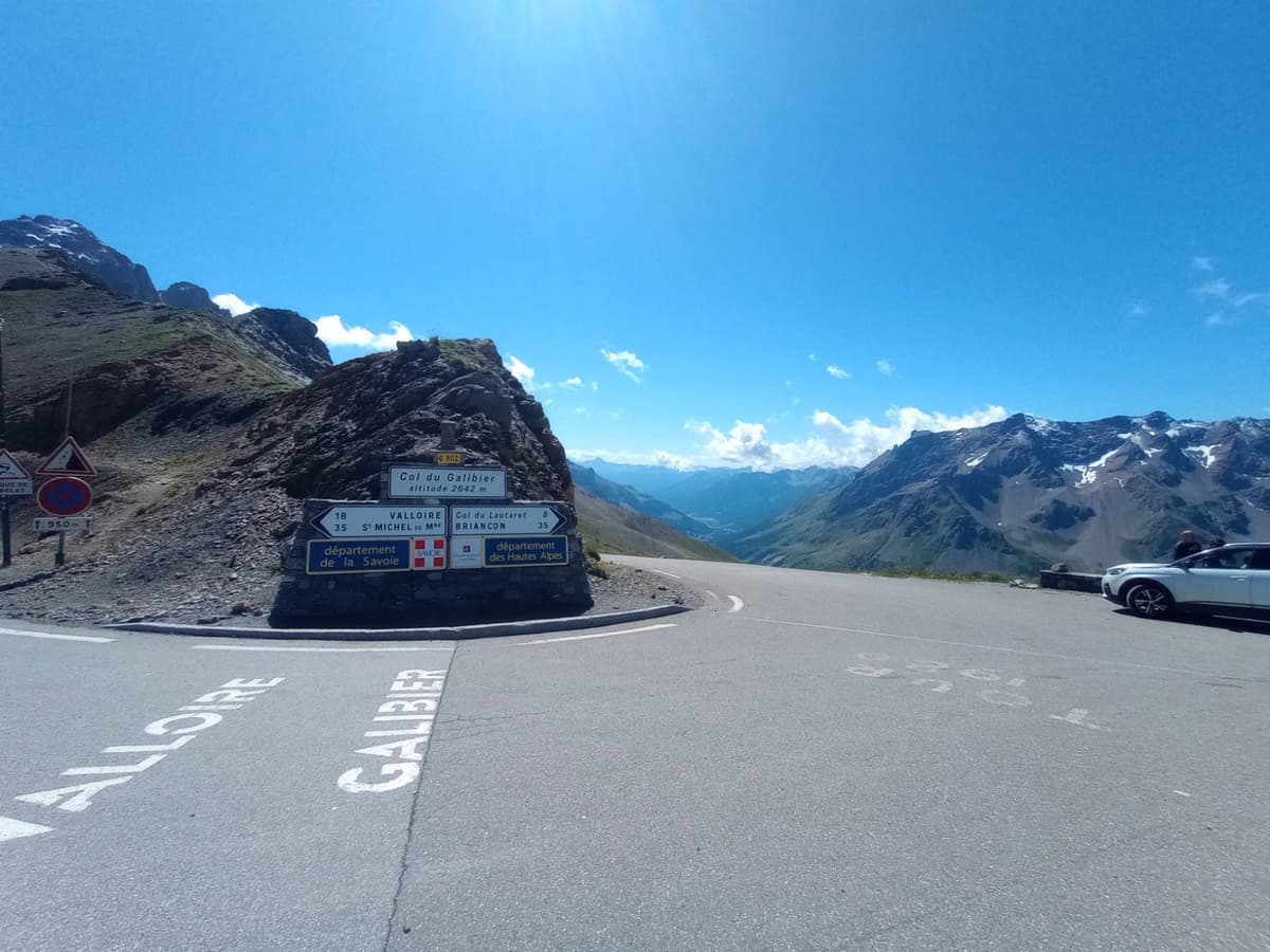 Een wegwijzer op de top van de Col du Galibier, 2.642 meter hoog, met op het asfalt de namen Valloire en Galibier geschilderd en een zicht op besneeuwde bergen.