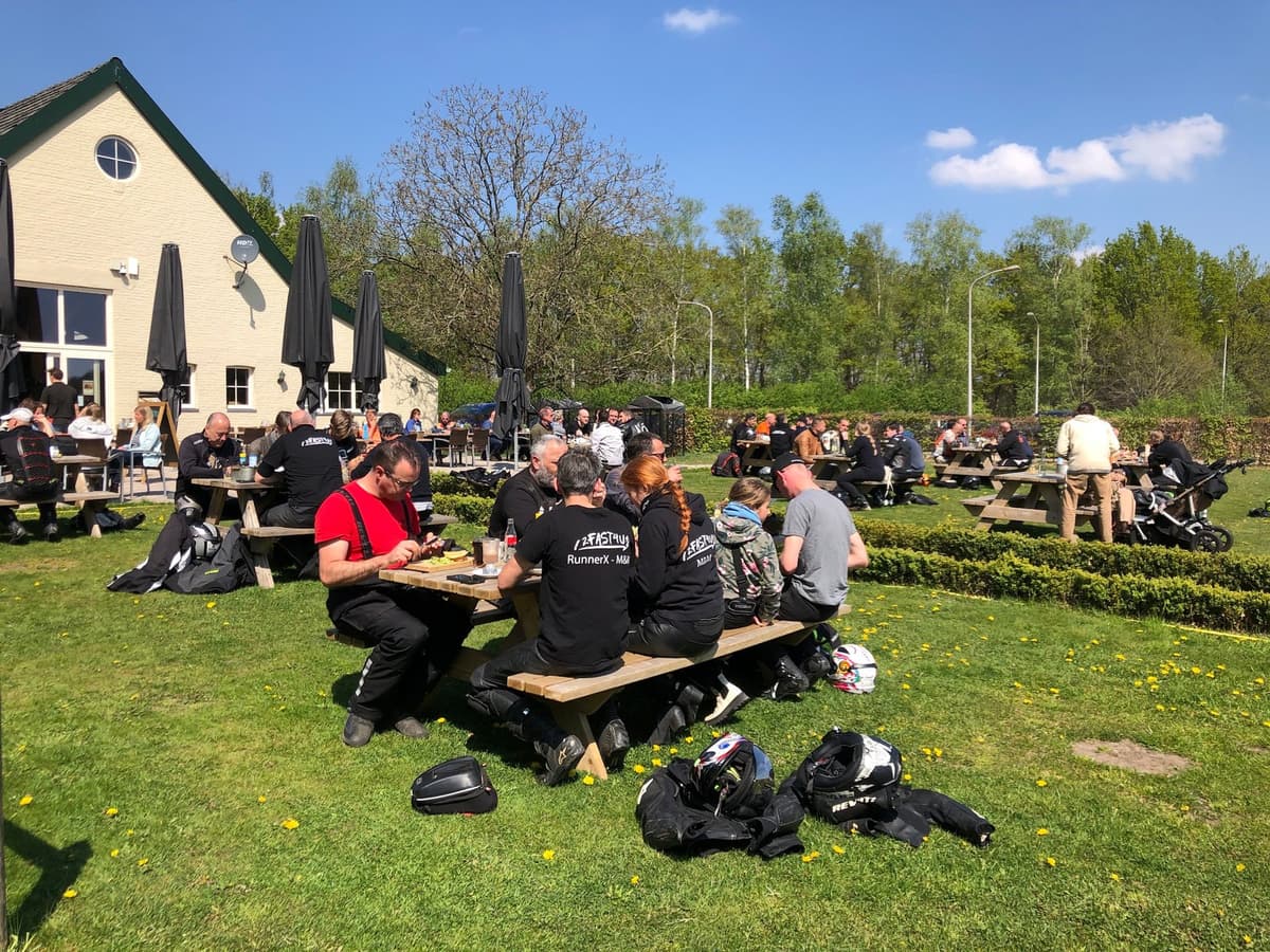 Motorrijders zitten aan houten picknicktafels op een grasveld in de zon, met helmen en jassen in het gras, voor een gebouw met parasols.