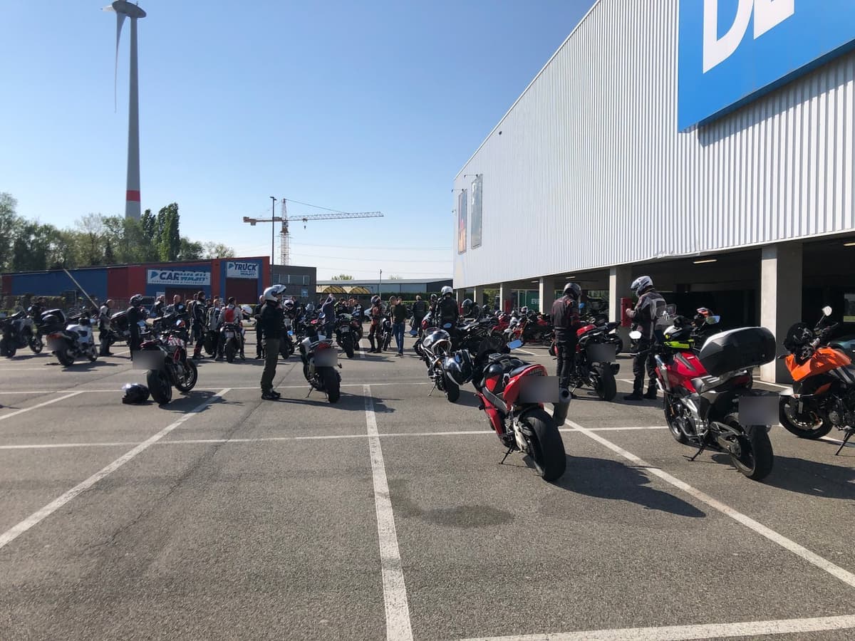 Tientallen motoren en motorrijders op een grote parking voor een winkelgebouw, onder een strakblauwe lucht met een windmolen.