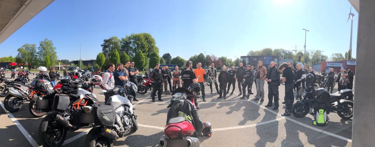 Een grote groep motorrijders staat in een kring rond de briefing op een zonnige parking, tussen tientallen geparkeerde motoren.