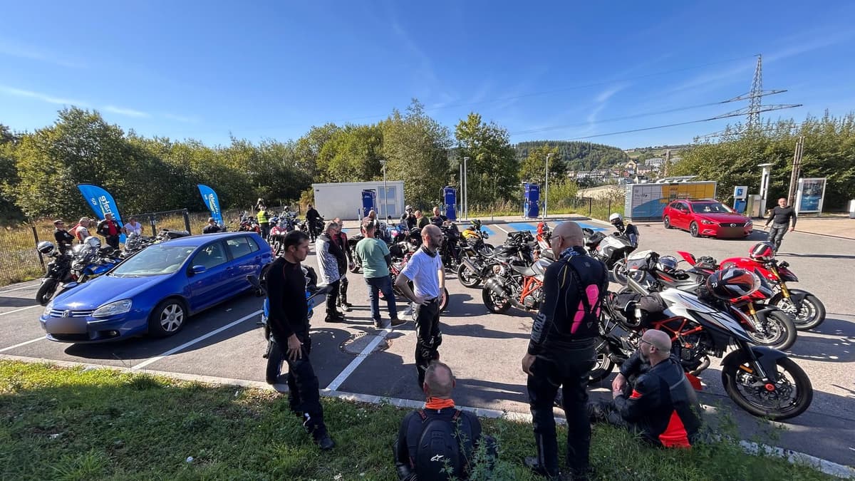 Tientallen motoren en motorrijders op de parking van een tankstation in de zon, met beboste heuvels en een dorpje op de achtergrond.