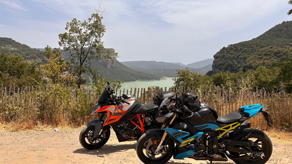 Een oranje KTM en een zwarte BMW staan op een grindpad boven een turquoise stuwmeer, tussen beboste hellingen en rotswanden.
