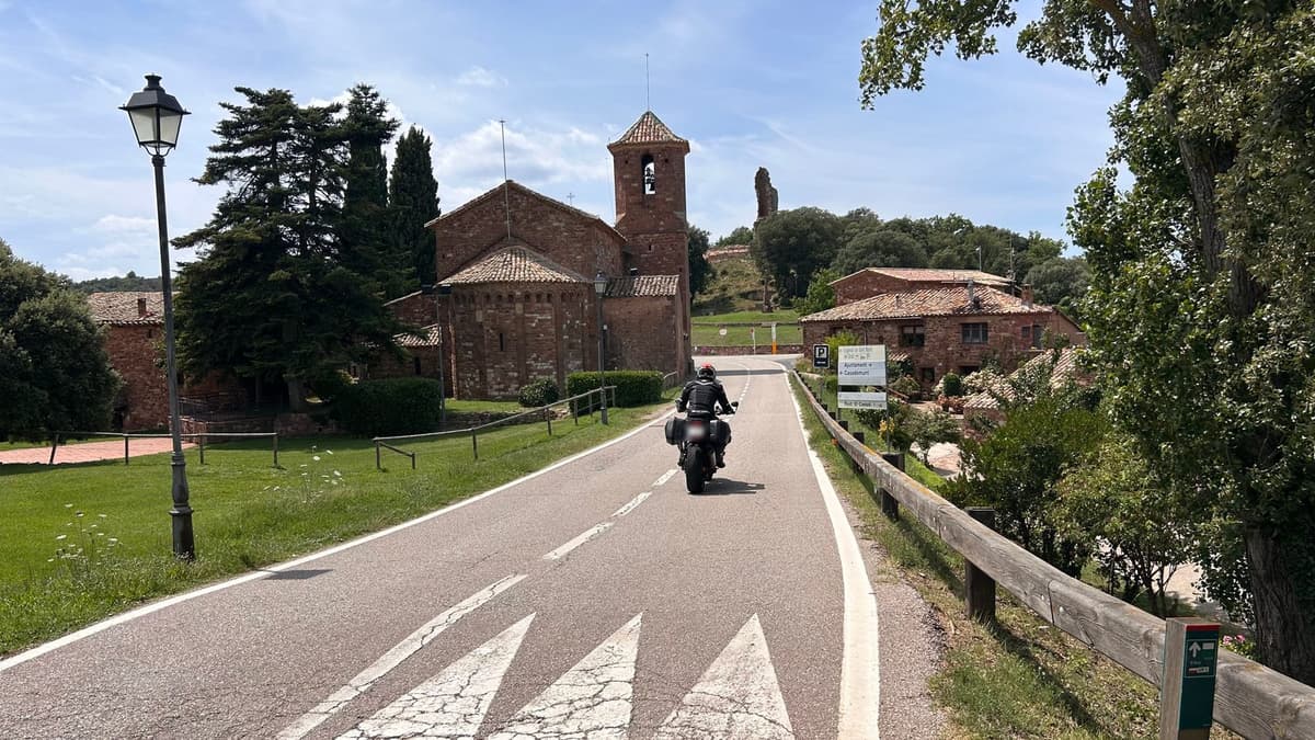 Een motorrijder met zijkoffers rijdt over een smalle weg een dorpje binnen, langs een romaans kerkje van rode steen met een vierkante klokkentoren.
