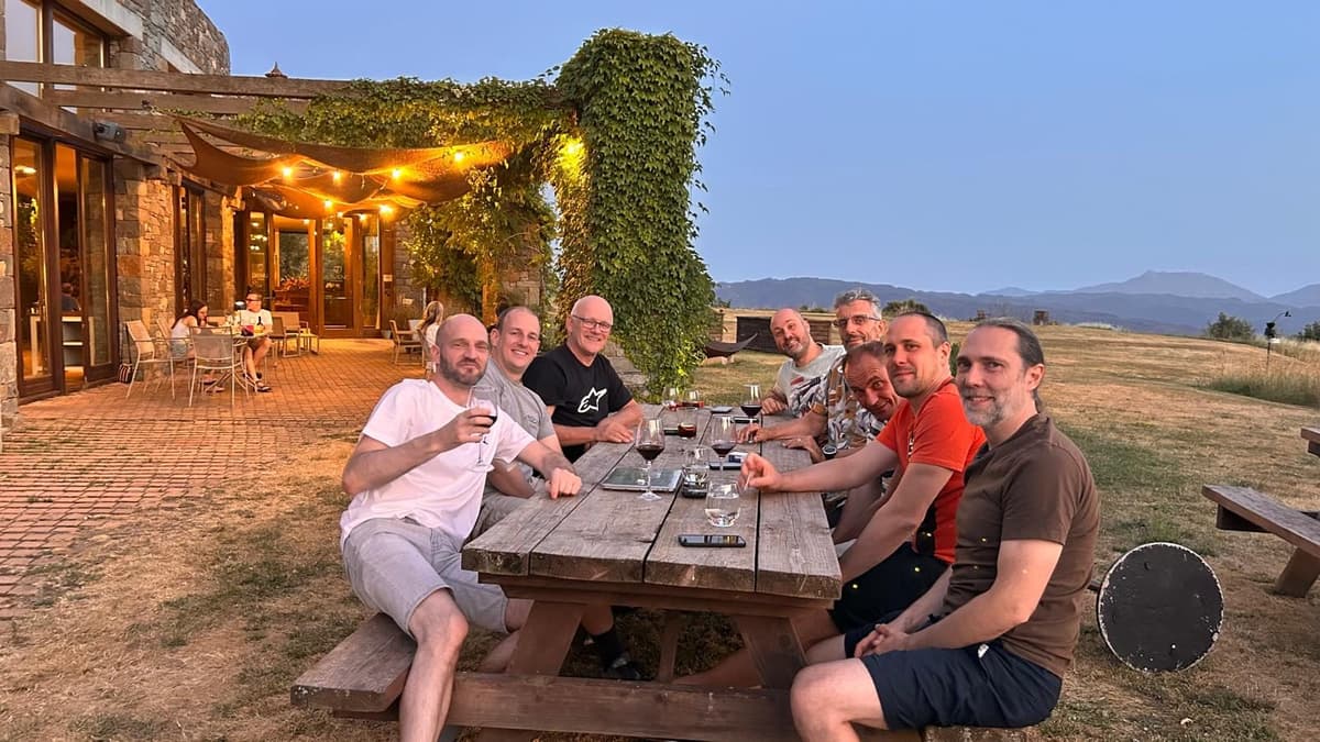 Een groep motorrijders zit 's avonds aan een houten picknicktafel buiten bij het hotel, met glazen rode wijn, lichtjes onder een pergola vol klimop en bergen aan de horizon.