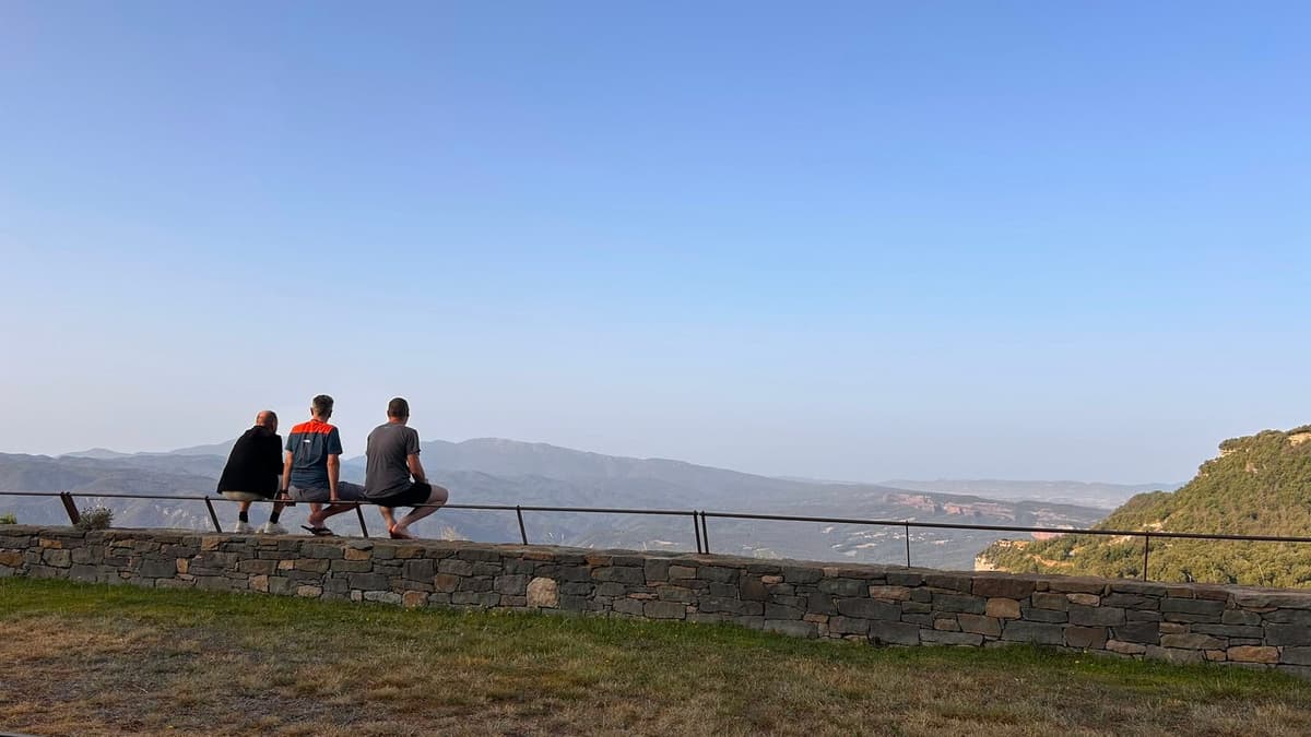 Drie mannen zitten met de rug naar de camera op een stenen muur met een reling en kijken uit over een wijd dal met bergen.