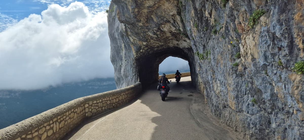 Twee motorrijders rijden op een smalle weg door een tunnel in een rotswand, met naast de weg een zee van wolken.