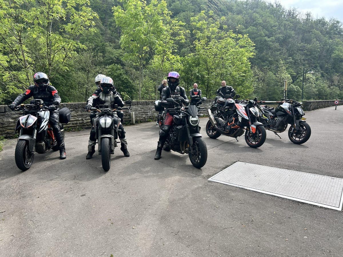 Motorrijders met helm op zitten op hun naked bikes op een parking tussen beboste hellingen, met daarachter nog een paar rijders bij hun motoren.