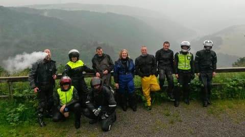 Een groep motorrijders in regenpakken poseert bij een houten vangrail, met achter hen een vallei in de mist.