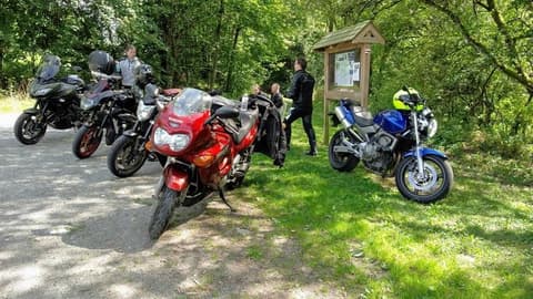Vijf motoren staan aan de rand van een bos naast een houten infobord, waar een paar motorrijders even uitrusten.