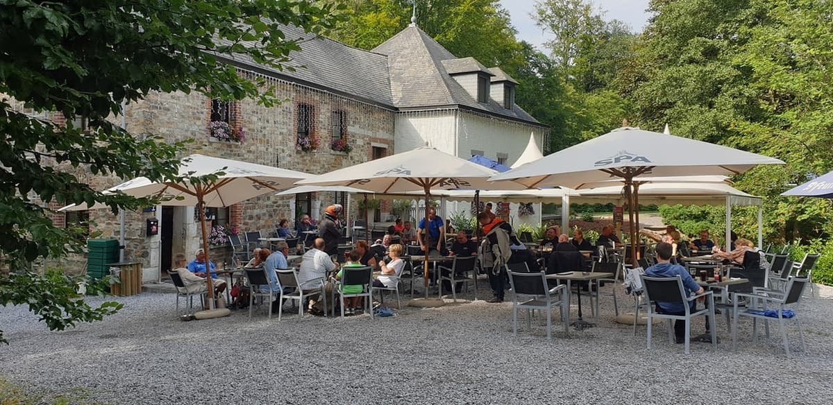 Een vol terras met witte parasols voor een oud stenen gebouw tussen de bomen, waar motorrijders aan tafel zitten en bijpraten.