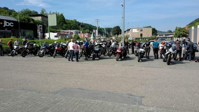 Een lange rij motoren en motorrijders op een parking onder een blauwe lucht, met beboste heuvels op de achtergrond.