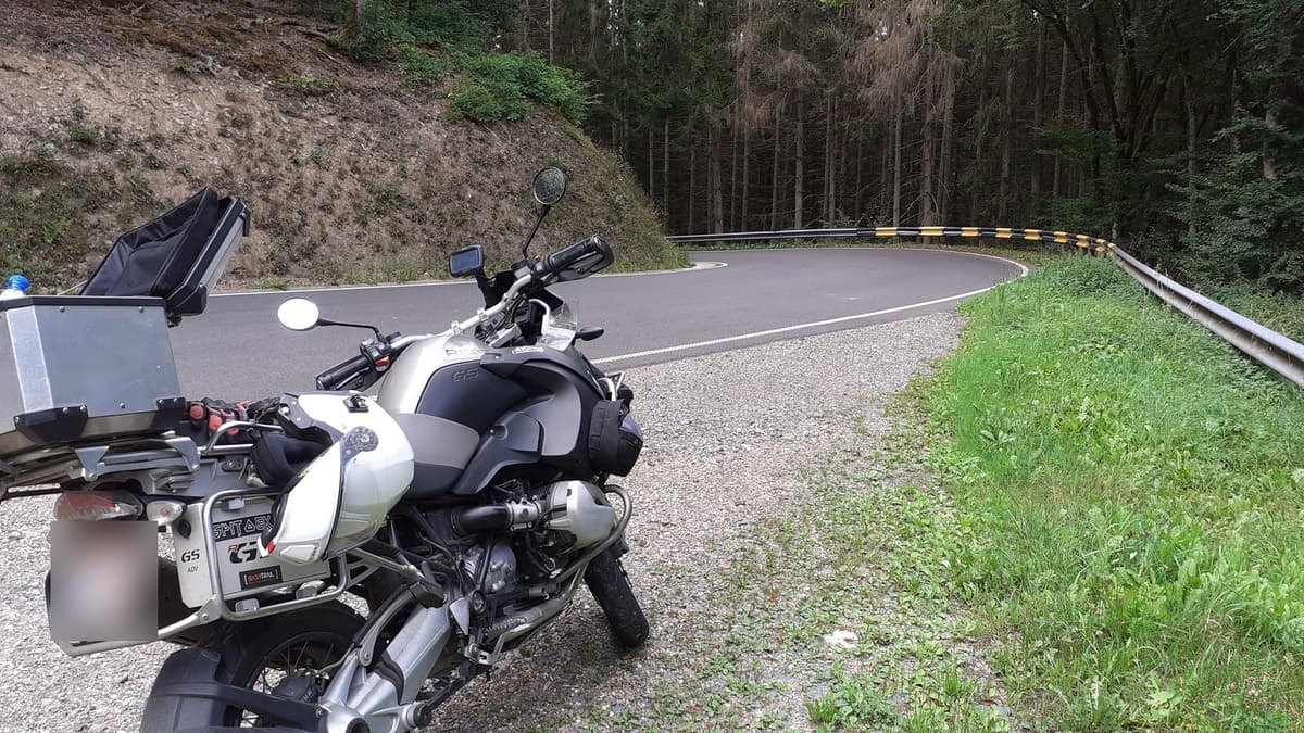 Een zilveren BMW GS met koffers in de berm, voor een haarspeldbocht met vangrail tussen de dennen.