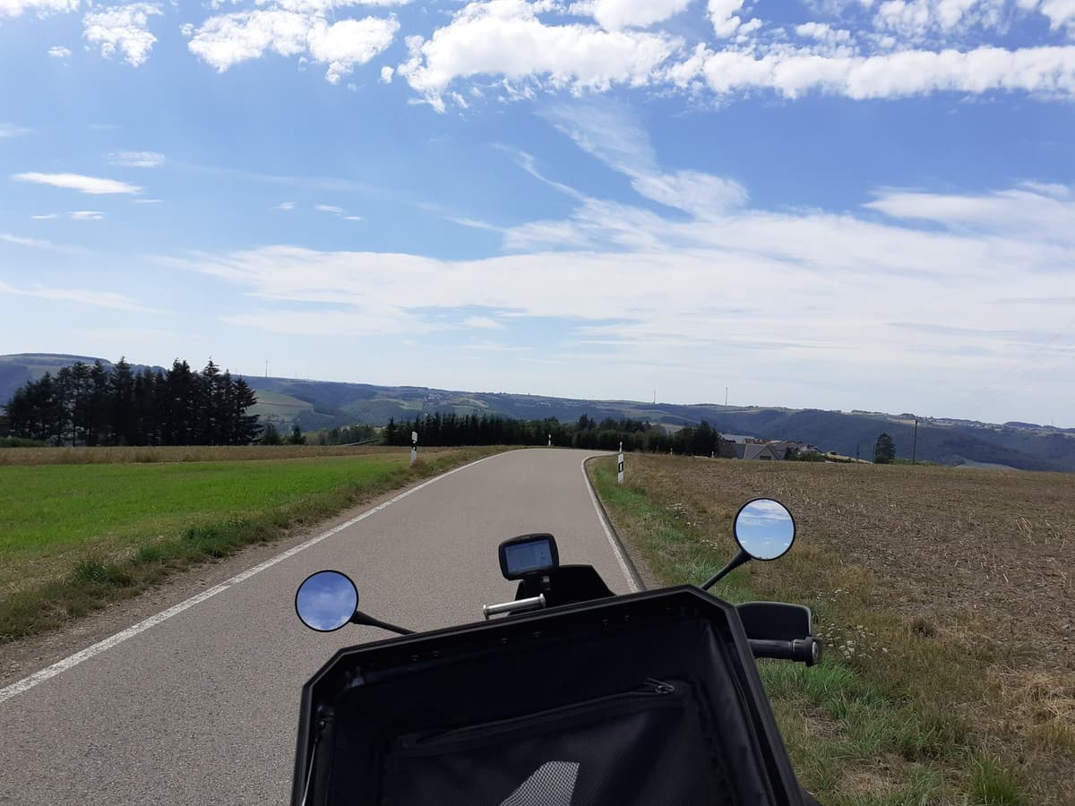 Zicht over het stuur en de tanktas van een motor op een smal baantje tussen de velden, met beboste heuvels en windmolens in de verte.