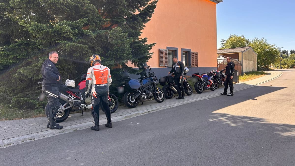 Vier motorrijders in motorpak staan lachend bij hun motoren, netjes op een rij geparkeerd langs een oranje huis en een grote spar, in de zon.
