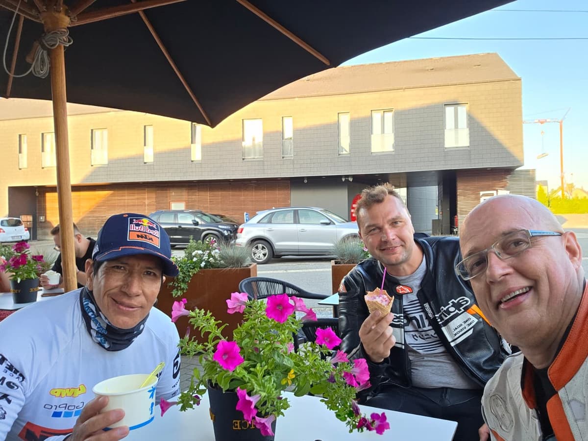Drie lachende motorrijders aan een tafeltje op een terras onder een parasol, twee met een ijsje, in het lage avondlicht.