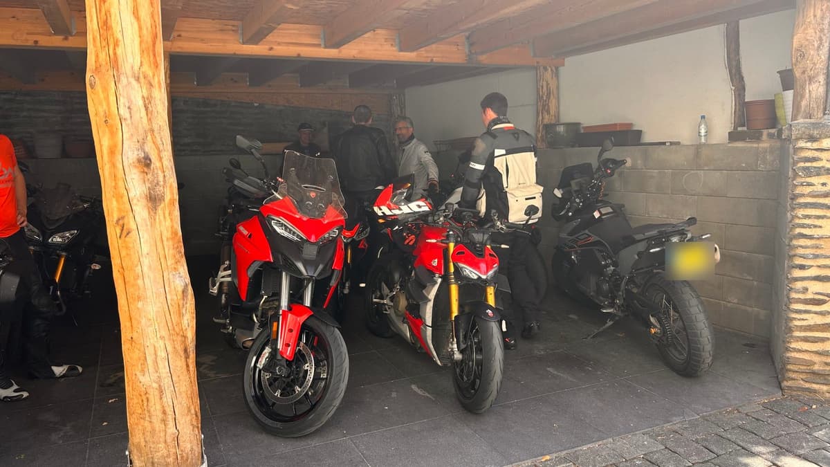 Motoren, onder meer twee rode Ducati's en een adventuremotor, staan onder een houten afdak, met erachter een paar motorrijders in gesprek.