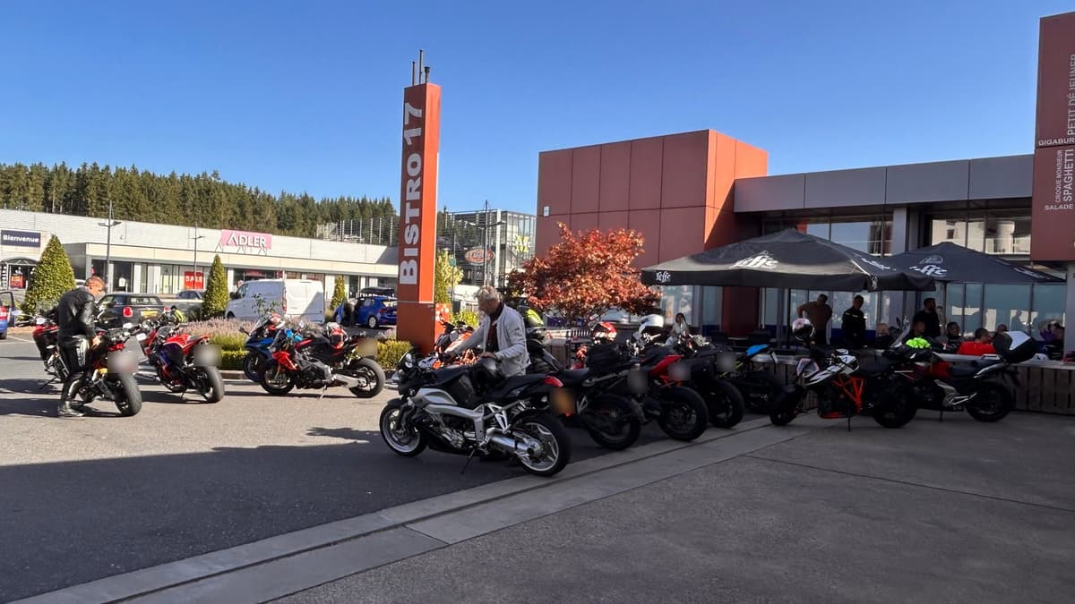 Een lange rij motoren, van sportmotoren tot een KTM en een BMW-toerfiets, op de parking van een tankstation onder een strakblauwe lucht, met een paar rijders bij hun motor en anderen op het terras.