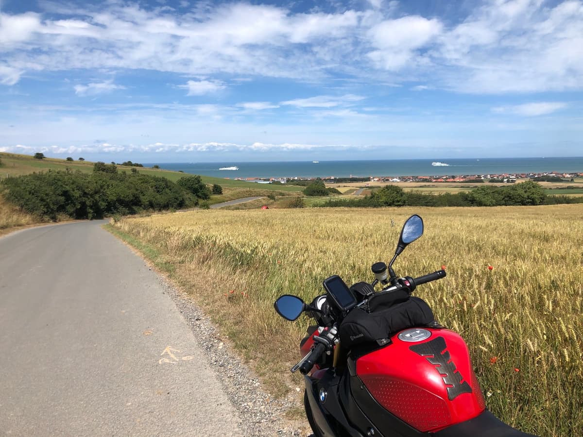 Zicht vanaf een rode motor op een smalle weg die tussen graanvelden afdaalt naar de kust, met de zee en witte ferry's aan de horizon.