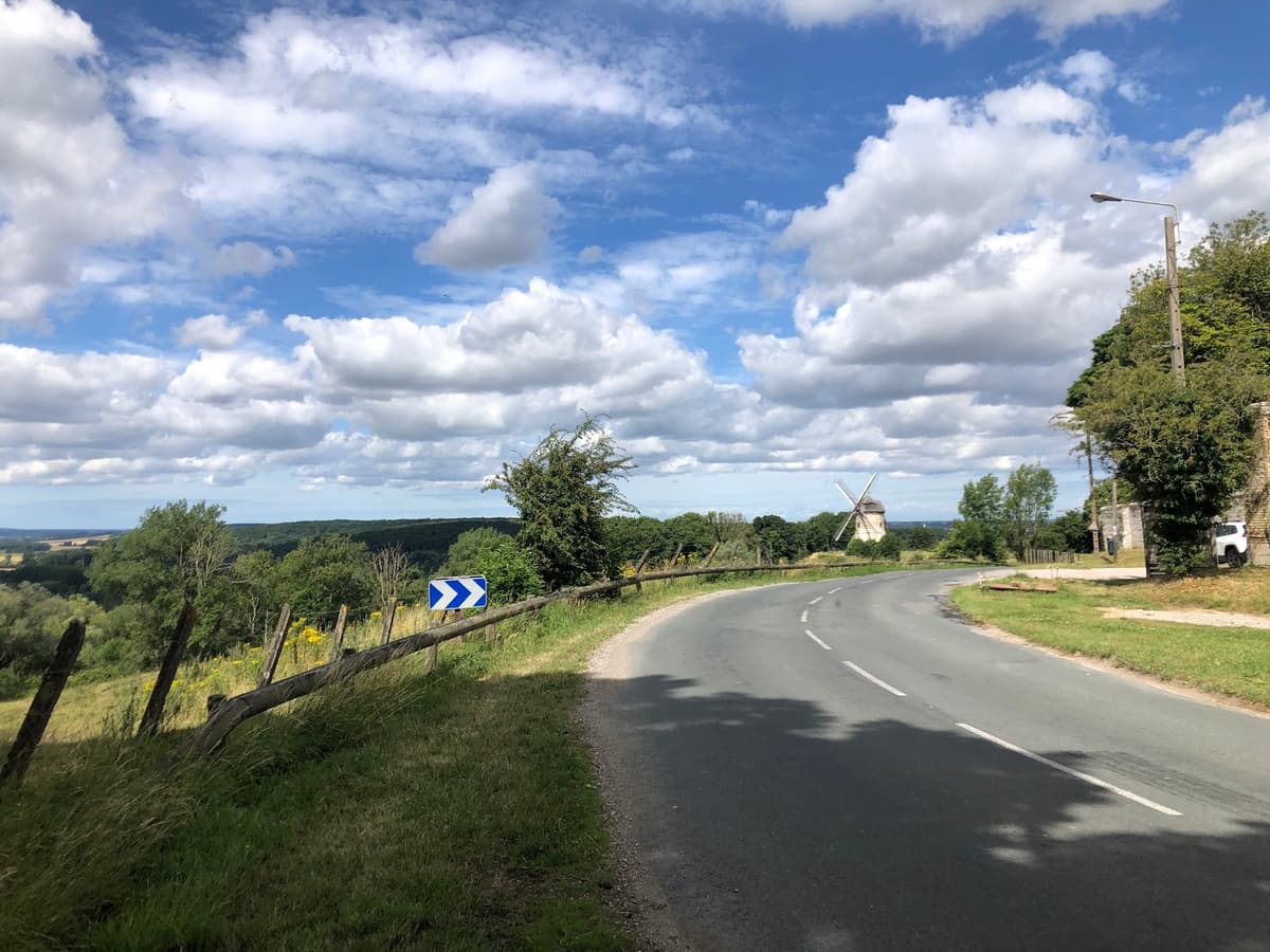 Een landweg die naar rechts draait langs een houten afsluiting en een wit-blauw bochtenbord, met een windmolen en een weids zicht over het heuvelland.