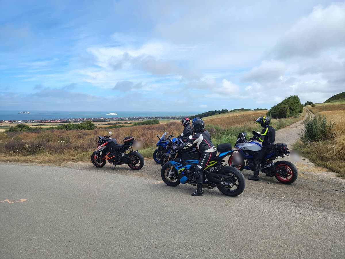 Vier motorrijders met hun motoren aan de rand van een weg op een heuvel, met beneden een kustdorp en de zee.