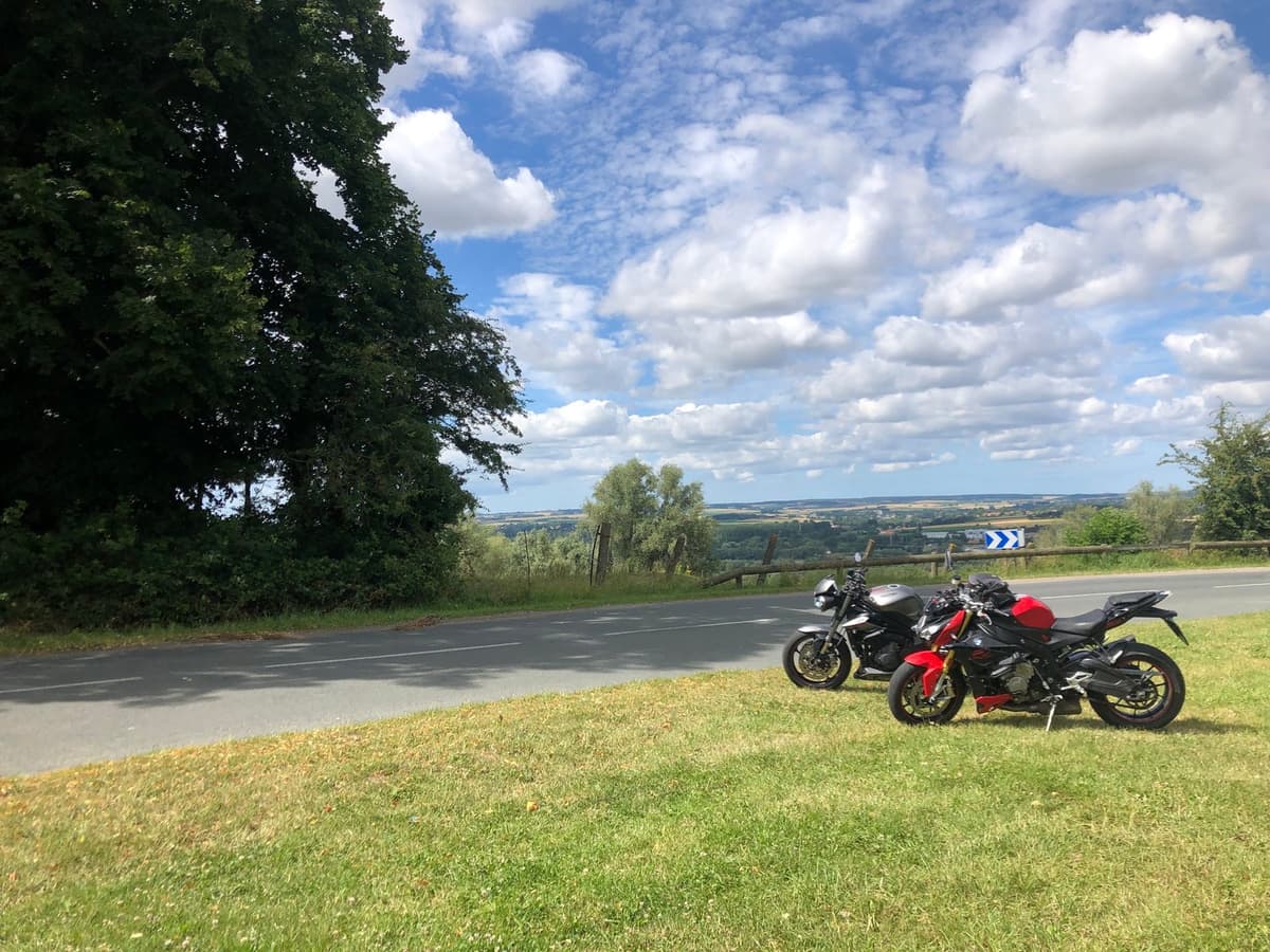 Twee naked bikes, een grijze en een rood-zwarte, op het gras langs een weg met een bochtenbord en uitzicht over het binnenland.