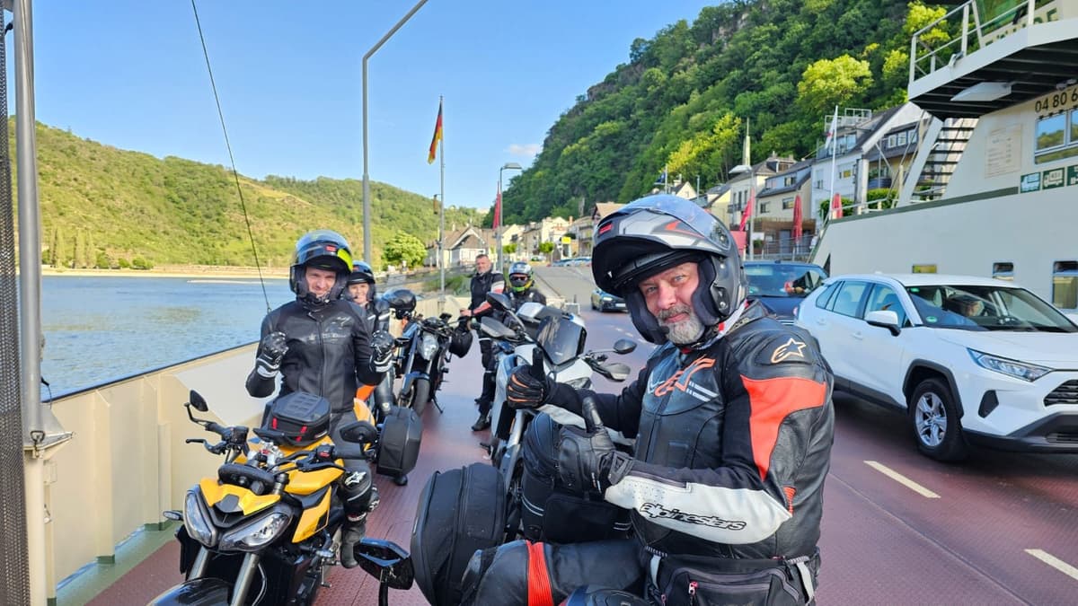 Lachende motorrijders met helm op bij hun motoren op een veerboot over de Rijn, met een duim omhoog, tussen auto's en een dorp onder een beboste helling.