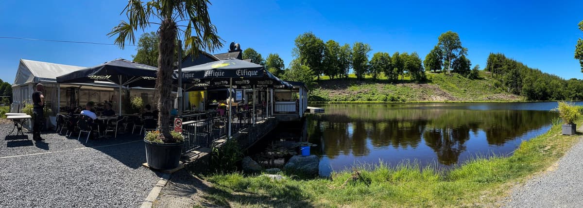 Een terras met zwarte parasols en een palmboom aan een vijver, met beboste heuvels en een blauwe lucht.