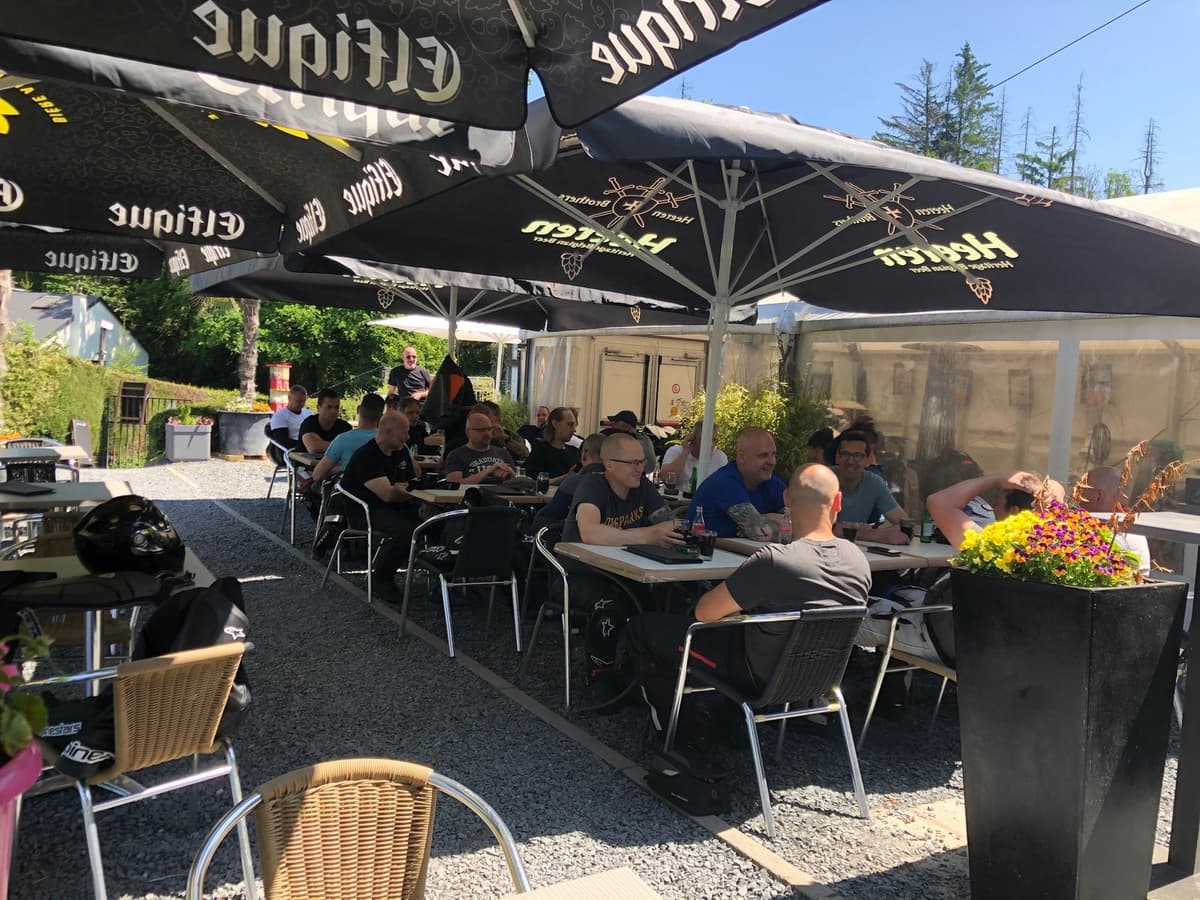 Motorrijders aan tafels onder zwarte parasols op een zonnig terras.
