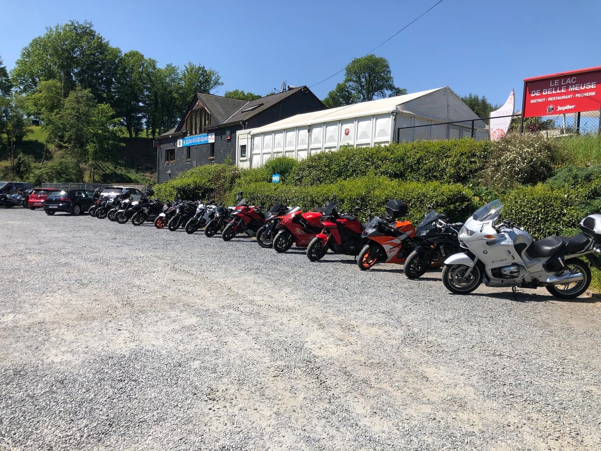 Een lange rij motoren op een grindparking voor een bistro en een witte feesttent, met het rode bord van Le Lac de Belle Meuse.