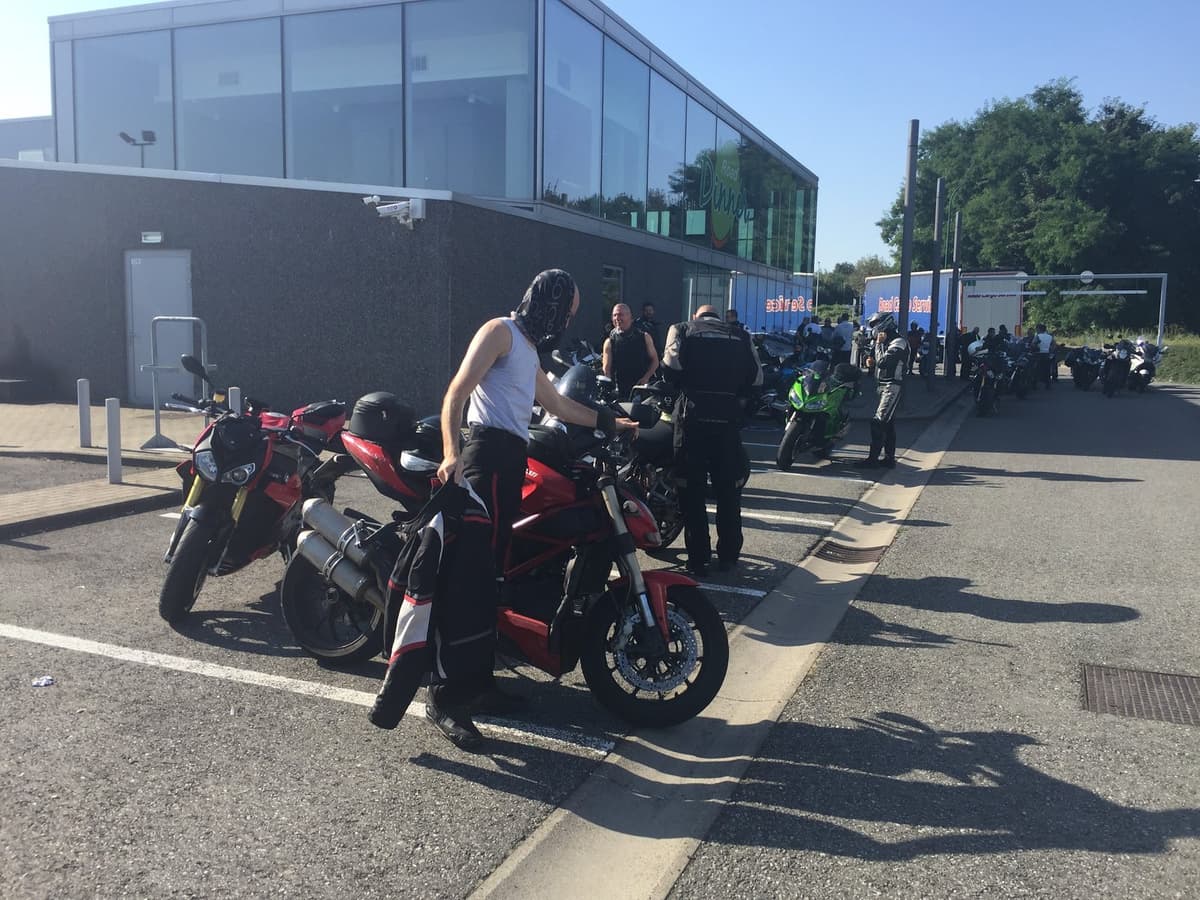 Een motorrijder stapt op een rode Ducati op de parking van een tankstation, tussen andere motoren en rijders.