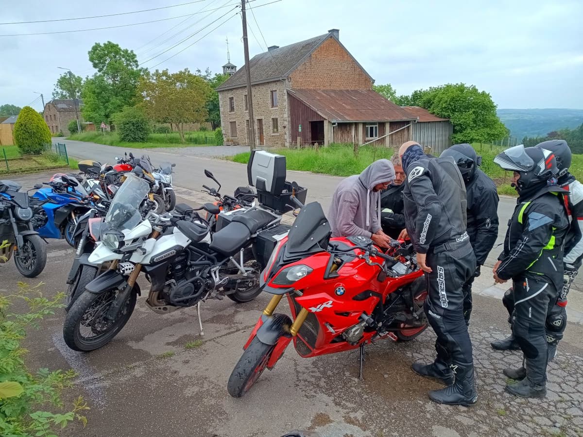 Motorrijders staan rond een rode BMW tussen geparkeerde motoren, voor een oude stenen boerderij.